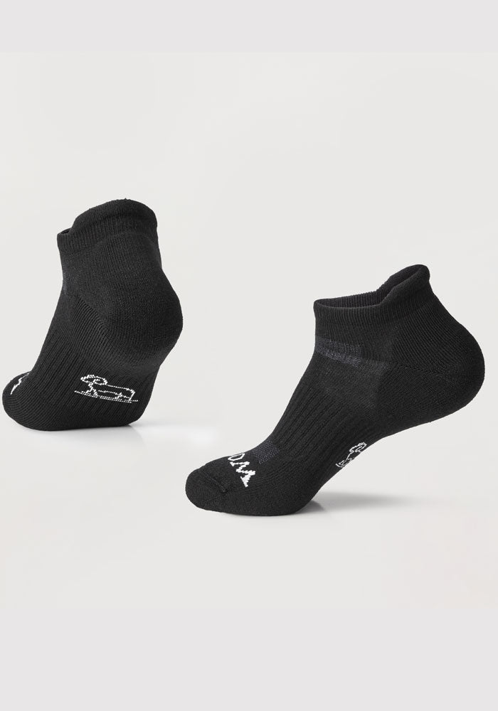 Two black Rowan Cushioned Ankle Socks by Woolx are displayed against a white background. One sock is positioned upright, showing the top and heel, while the other is placed at an angle to emphasize its side and sole. Both lightweight merino wool socks have small, discreet text embroidered near the toes.