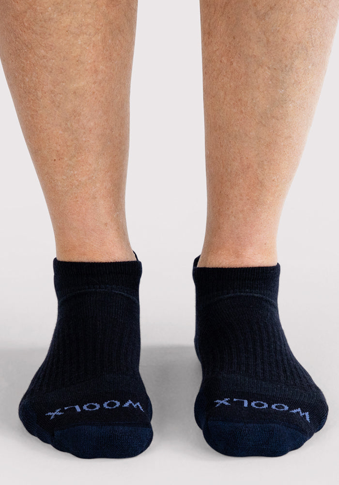 A person wearing Woolx Mens Rowan No Show Socks in Deep Night, featuring "WOOLX" in white on the toes, stands on a light gray surface.
