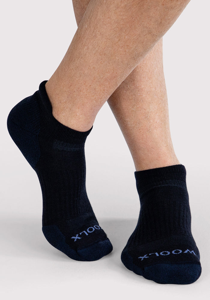 A close-up of a person wearing Woolx Mens Rowan No Show Socks in Deep Night, standing on a light gray surface with one foot crossed over the other. The black Merino wool socks feature white text near the toes.