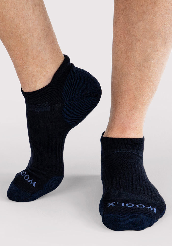 A close-up of a person wearing Mens Rowan No Show Socks in Deep Night by Woolx, featuring "Woolx" on the toes and standing on a light gray background.