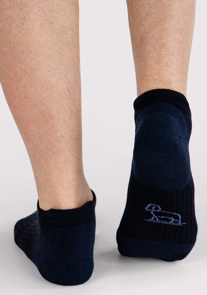 A close-up of a person's lower legs wearing Woolx Mens Rowan No Show Socks in Deep Night. The right sock displays a small light blue dachshund outline near the heel.