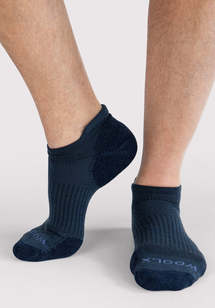 A person stands on tiptoe, one foot slightly raised, showcasing the ribbed texture and snug fit of Woolx Mens Rowan No Show Socks in Evening Tide against a light background.