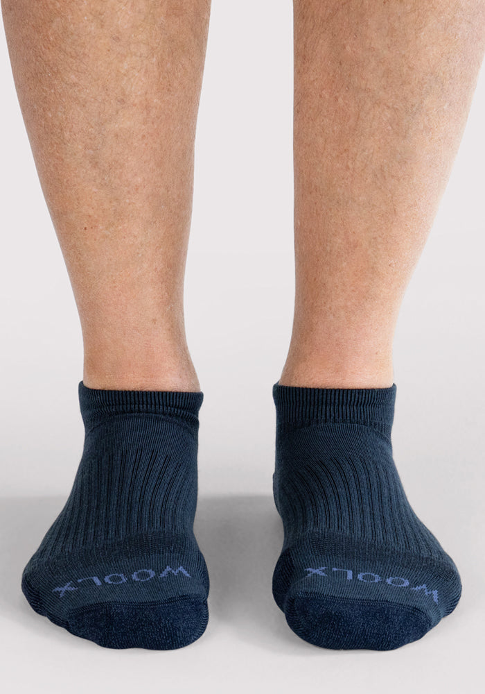 A pair of feet wearing Woolx Mens Rowan No Show Socks in Evening Tide, with "WOOLX" on the toes, standing on a light grey surface.