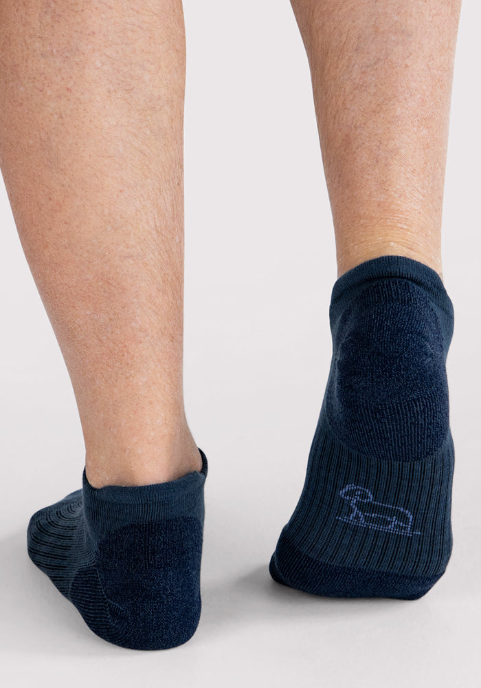 Back view of a person's lower legs and feet wearing Woolx Mens Rowan No Show Socks in Evening Tide, featuring cushioned dark blue ankle socks with a small dog outline, standing on a light surface.