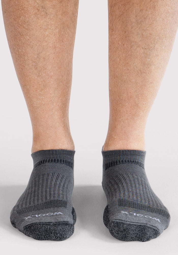 A close-up of a person’s lower legs and feet wearing Woolx Mens Rowan No Show Socks in grey, featuring a ribbed texture, dark toe areas, and natural moisture-wicking comfort, standing on a light-colored surface.