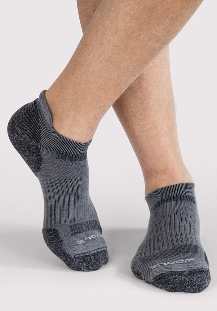 A person with crossed legs wears Woolx Mens Rowan No Show Socks in grey, featuring darker heels and toes, against a light background. The socks are moisture-wicking, comfortable, and ankle length.