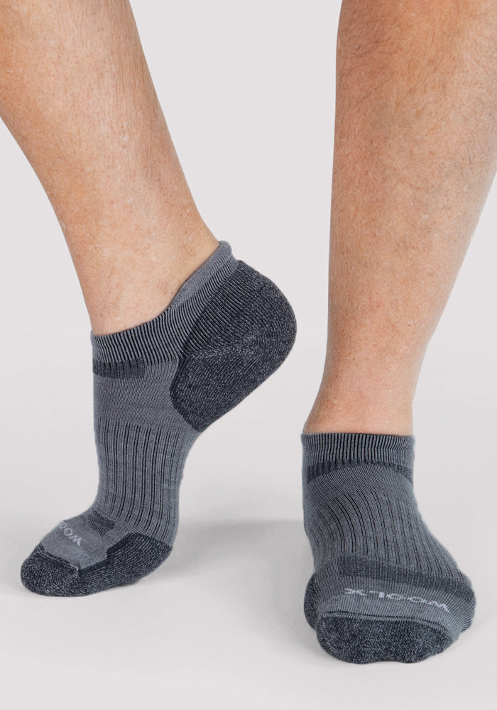 Close-up of a person’s lower legs wearing Woolx Mens Rowan No Show Socks in grey, featuring dark gray accents, standing on a light background with one heel slightly raised.