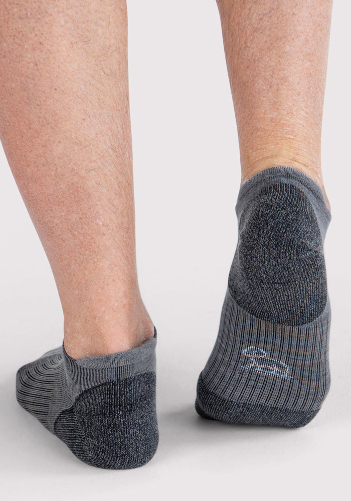 A close-up of a person wearing Woolx Mens Rowan No Show Socks in grey, featuring cushioned heels, textured bottoms, and moisture-wicking fabric as they stand on a light-colored surface.
