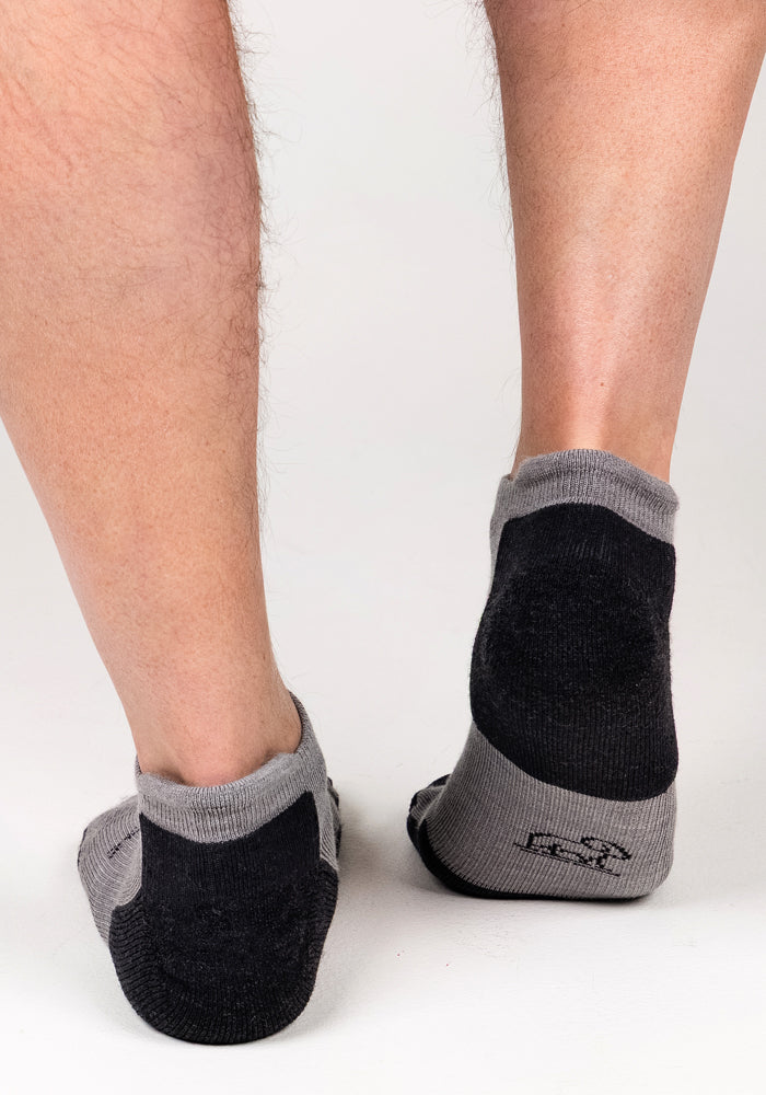 A close-up of the back of a person's lower legs and feet wearing Woolx Mens Rowan No Show Socks in Pebble, standing on a white surface.
