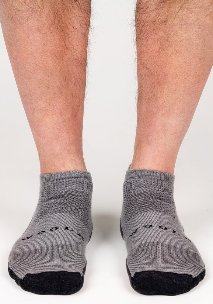 A close-up of a person's lower legs and feet wearing Woolx Mens Rowan No Show Socks in Pebble with black toes and soles, standing on a white surface.