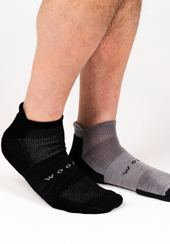 A person wearing Woolx Mens Rowan No Show Socks in Pebble, one black and one gray, stands on a white background. The textured socks feature small white text on each and offer all-day comfort.