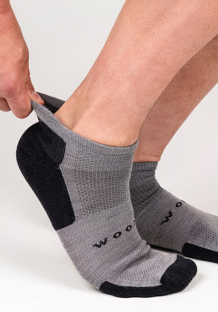A person is pulling on a pair of Woolx Mens Rowan No Show Socks in Pebble, which feature a ribbed texture, moisture-wicking fabric, cushioned ankle support, and subtle text on top.