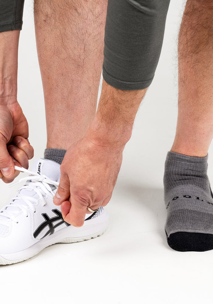 A person ties the laces of a white sneaker on one foot while wearing Woolx Mens Rowan No Show Socks in Pebble on the other, both feet resting on a white surface.
