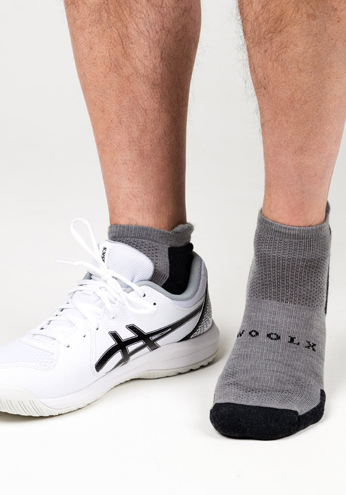 Close-up of a person’s lower legs wearing Woolx Mens Rowan No Show Socks in Pebble; one foot is in a white athletic shoe with black accents, the other wears just the sock, all set against a white background.