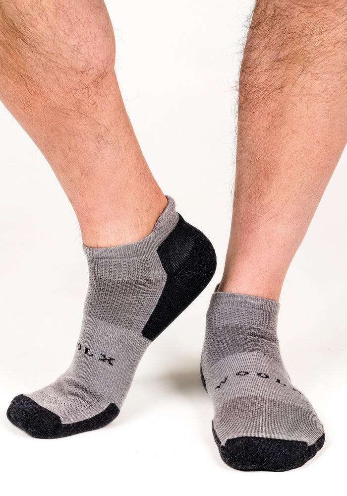 A person wears Woolx Mens Rowan No Show Socks in Pebble, featuring black soles and toes, text detail, textured uppers and heels, and moisture-wicking comfort, standing on a white background.