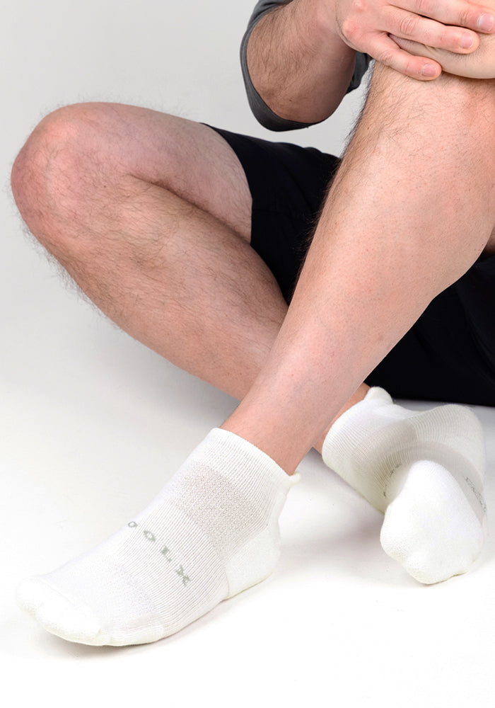 A person in black shorts and a gray top sits on the floor, highlighting their legs and feet in Woolx Mens Rowan No Show Socks - White Lightning. The cushioned socks feature green text, and the background is plain white.