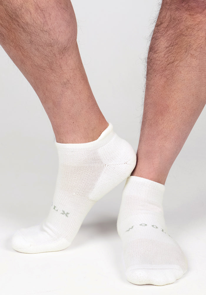A close-up of a person wearing Woolx Mens Rowan No Show Socks - White Lightning, standing on a white surface with feet crossed at the ankles. The white socks feature subtle green text near the toes.