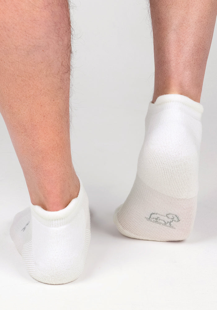 A close-up of two feet in Woolx Mens Rowan No Show Socks - White Lightning, standing on a white surface. These cushioned ankle socks have a subtle gray logo on the sole for a clean, modern look.