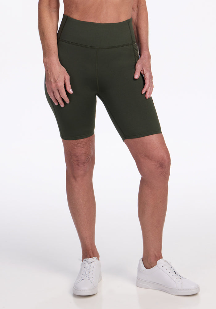 A person wearing Woolx Merino Flex® Dani Bike Shorts in Everleaf and white sneakers stands against a plain white background, with only the lower torso and legs visible. | Tam is 5’8.5”, wearing a size S