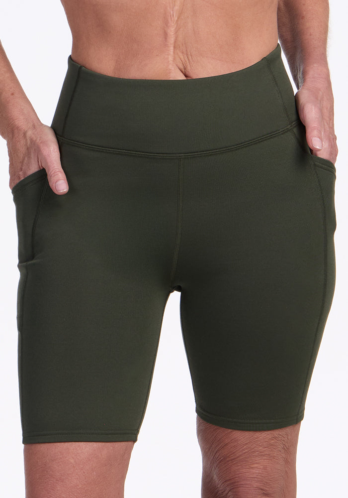 A person wears Woolx Merino Flex® Dani Bike Shorts in Everleaf, high-waisted and dark green with side pockets, standing with hands in the pockets. Only the lower torso and upper thighs are visible.