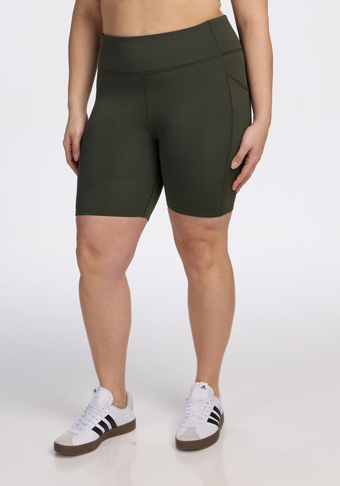 A person wearing Woolx Merino Flex® Dani Bike Shorts in Everleaf and white Adidas sneakers with black stripes stands against a plain white background, with their face not visible. | Juliana is 5'10", wearing a size XL