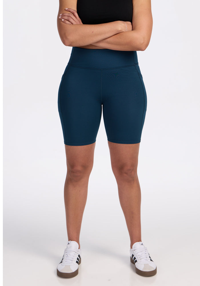 A person stands with arms crossed in a black sleeveless top, white sneakers with black stripes, and Woolx Merino Flex® Dani Bike Shorts in Majolica Blue, stylish moisture-wicking activewear set against a plain white background. | Kisha is 5’8”, wearing a size M
