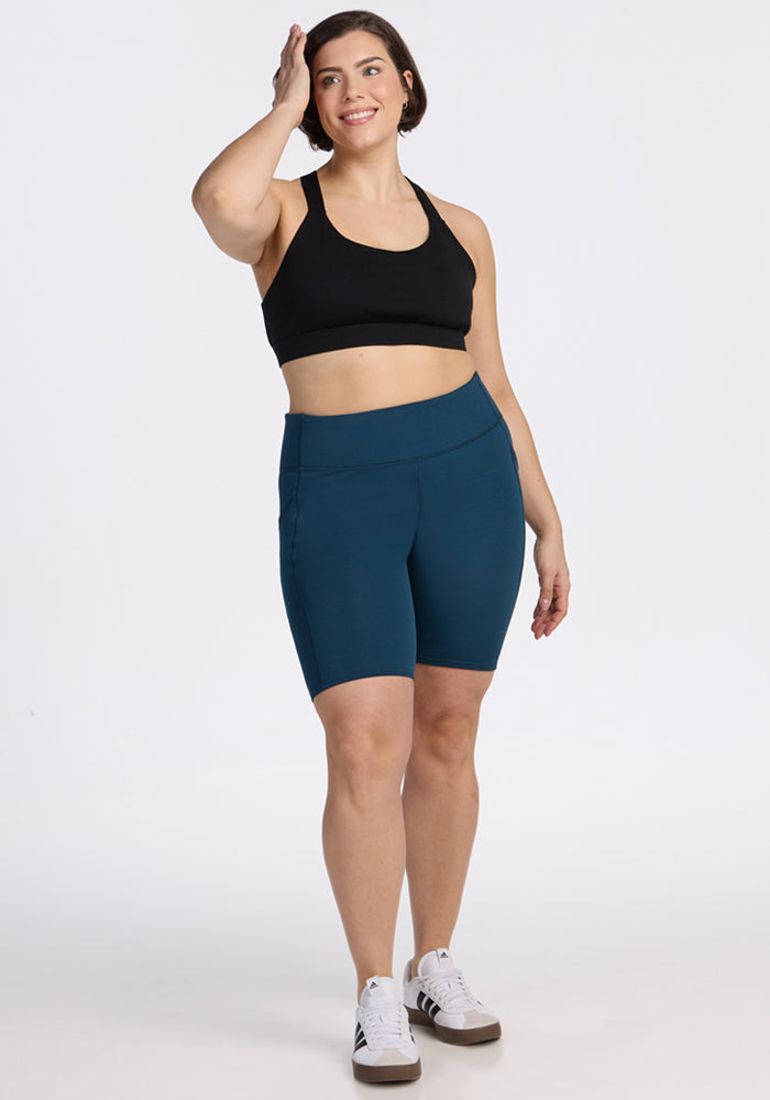 A woman in a black sports bra, Woolx Merino Flex® Dani Bike Shorts in Majolica Blue, and white sneakers stands smiling with one hand touching her head against a plain white background.