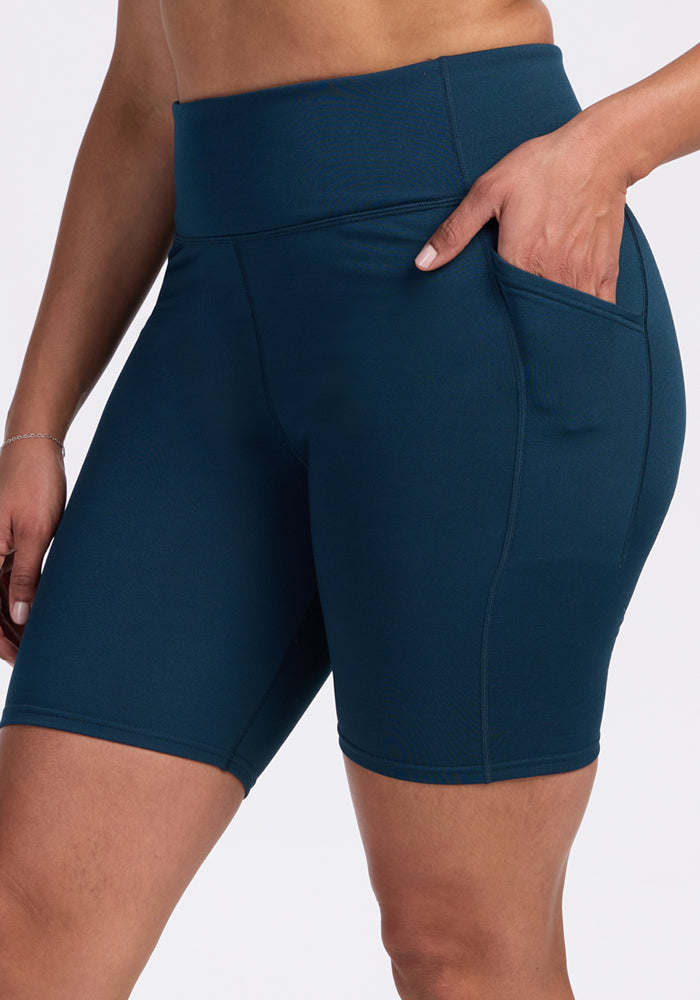 A person wearing Woolx Merino Flex® Dani Bike Shorts in Majolica Blue, with a high-waisted, fitted design and side pocket, places their hand in the pocket. The moisture-wicking activewear shorts are shown cropped at the torso and mid-thigh.
