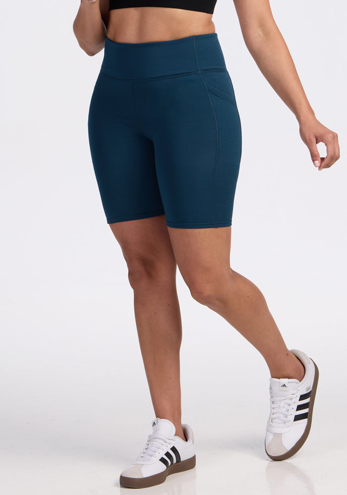 A person is standing against a plain white background, cropped from the shoulders up, wearing Woolx Merino Flex® Dani Bike Shorts in Majolica Blue, a black crop top, and white Adidas sneakers with black stripes.