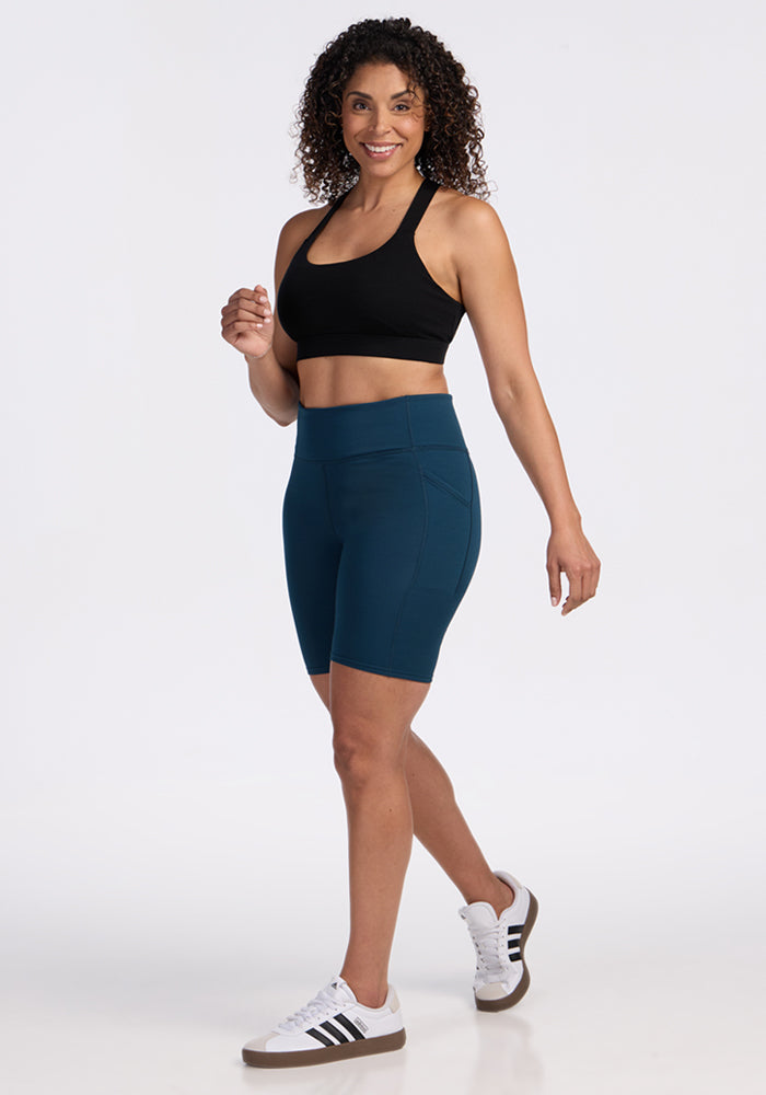 A woman with curly hair smiles while posing in a black sports bra, Woolx Merino Flex® Dani Bike Shorts in Majolica Blue, and white sneakers with black stripes against a plain white background.