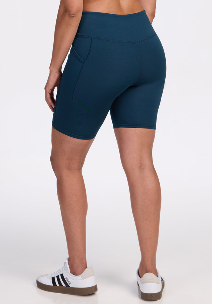 A person wears Woolx Merino Flex® Dani Bike Shorts in Majolica Blue, paired with white Adidas sneakers, standing facing away with arms relaxed at their sides against a plain white background.