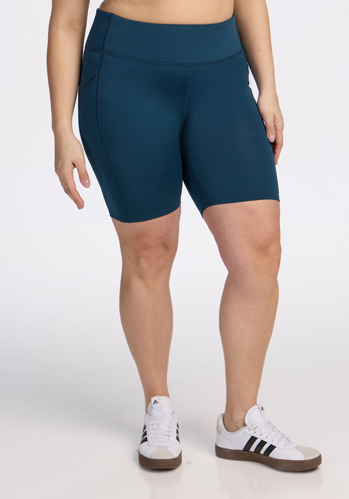 A person wearing Woolx Merino Flex® Dani Bike Shorts in Majolica Blue and white Adidas sneakers with black stripes stands against a plain white background. Their upper body is not visible. | Juliana is 5'10", wearing a size XL