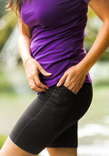 A person outdoors adjusts the moisture-wicking waistband of Woolx Merino Flex® Dani Bike Shorts - Black with one hand, while the other rests on the side pocket; they're wearing a fitted purple shirt against a backdrop of blurred greenery.