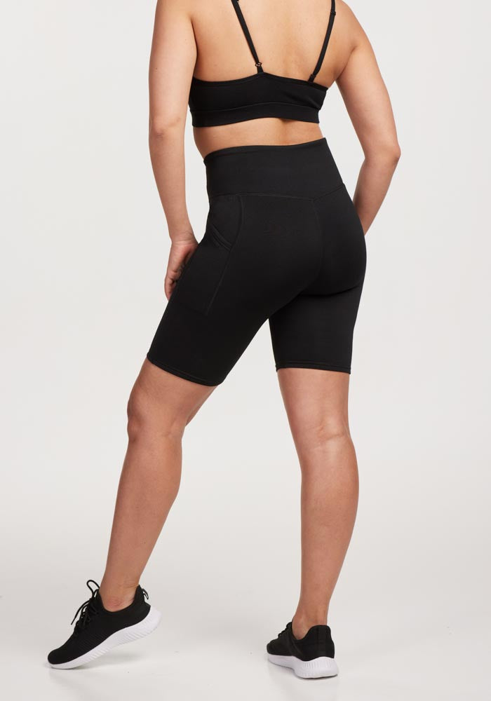 Someone wearing a black sports bra, Woolx Merino Flex® Dani Bike Shorts—recognized for their odor resistance—and black athletic shoes stands with their back to the camera against a plain white background.