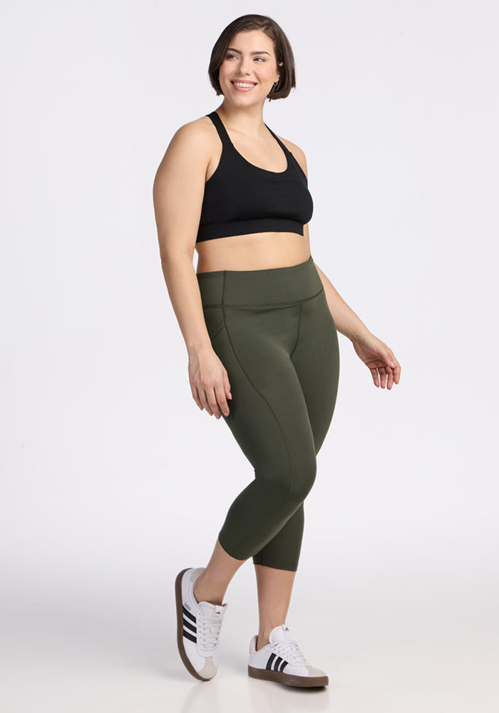 A woman with short brown hair smiles and poses in black sports bra, Woolx Merino Flex® McKenna Capris in Everleaf, and white Adidas sneakers against a plain white background.