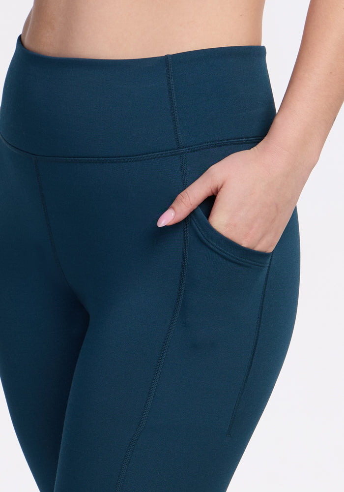 A person wears Woolx Merino Flex® McKenna Capris in Majolica Blue, shown from waist to mid-thigh with a hand in the side pocket against a white background. These capris provide moisture-wicking comfort for all-day wear.