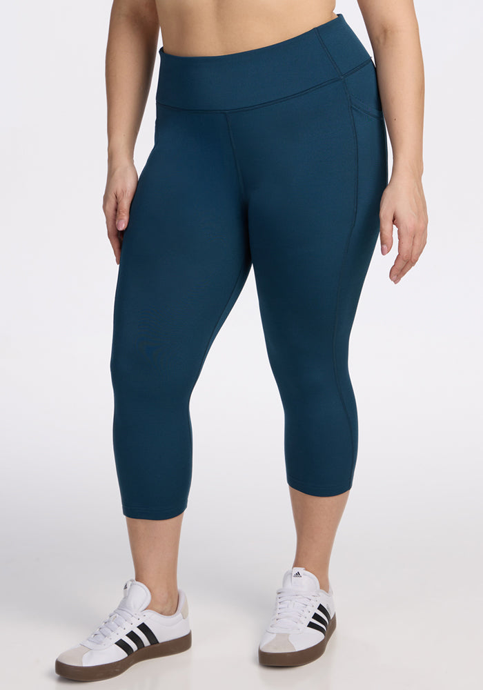 A person stands against a white background wearing Woolx Merino Flex® McKenna Capris in Majolica Blue and white Adidas sneakers with black stripes. The image is cropped waist to ankles. | Juliana is 5'10", wearing a size XL