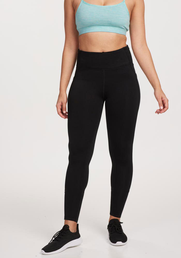 A person models Woolx Merino Flex® McKenna Petite leggings in black, paired with a light blue sports bra and black athletic shoes against a white background. The person's head is not visible in the image.