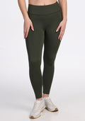 A person wearing Woolx Merino Flex® McKenna Petite - Everleaf high-waisted dark green leggings with pockets and white sneakers stands against a plain white background, showing only their lower body with hands resting on their thighs.