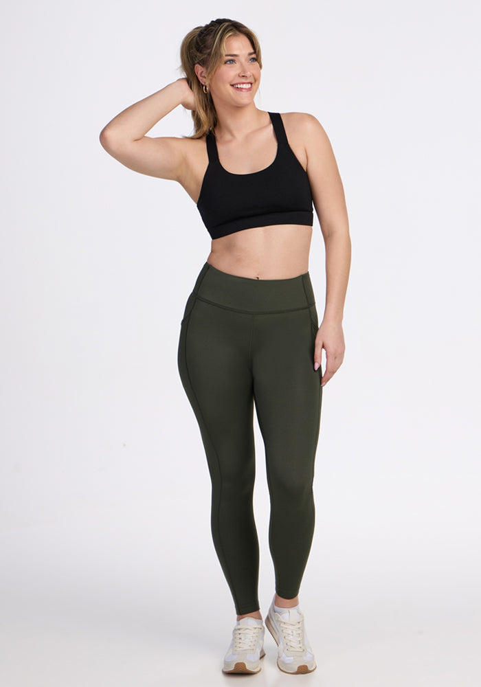 A woman smiles with one hand on her head, wearing Woolx Merino Flex® McKenna Petite - Everleaf olive green leggings with pockets, a black sports bra, and white sneakers against a plain white background.