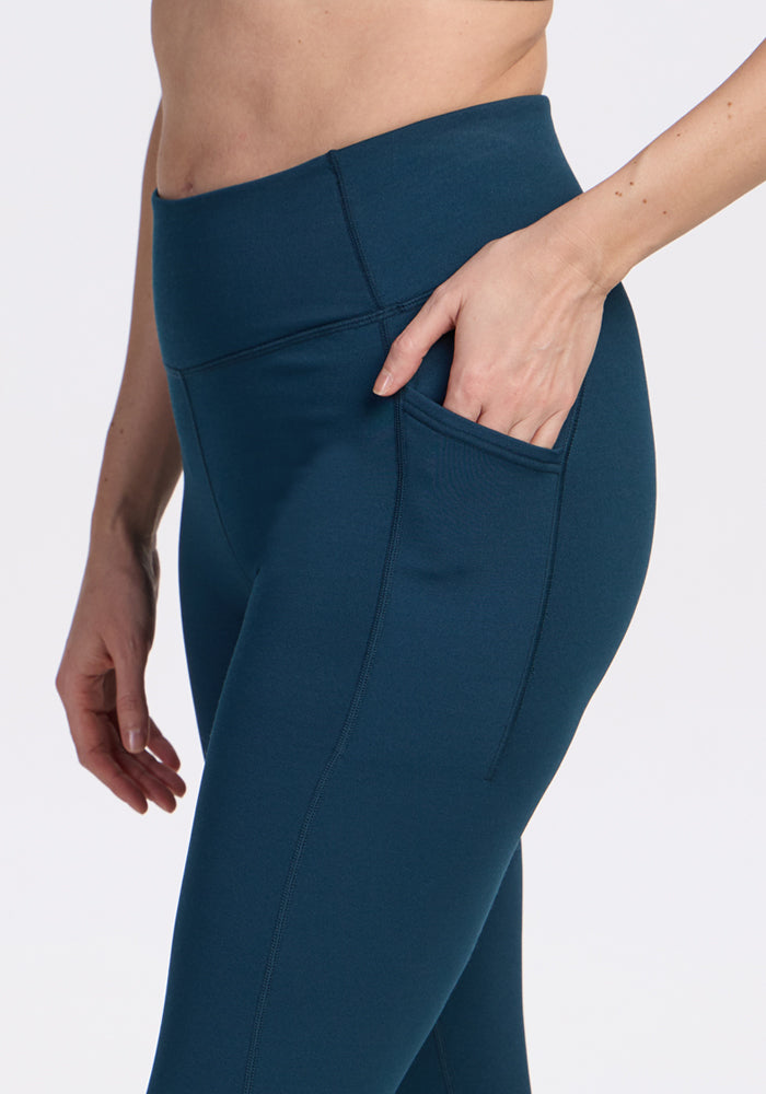 A person wears Woolx Merino Flex® McKenna Petite leggings in Majolica Blue, shown waist to mid-thigh with a hand in the side pocket, against a plain light background.