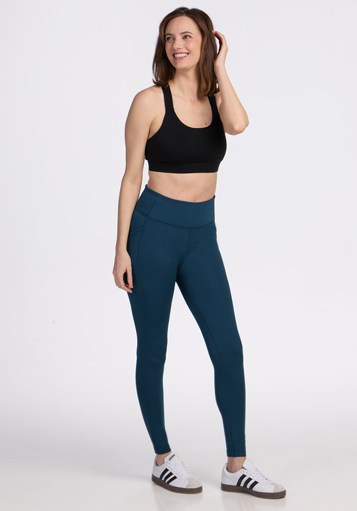 A woman stands smiling in a black sports bra, Woolx Merino Flex® McKenna Petite leggings in Majolica Blue, and white sneakers with black stripes, set against a plain white background.