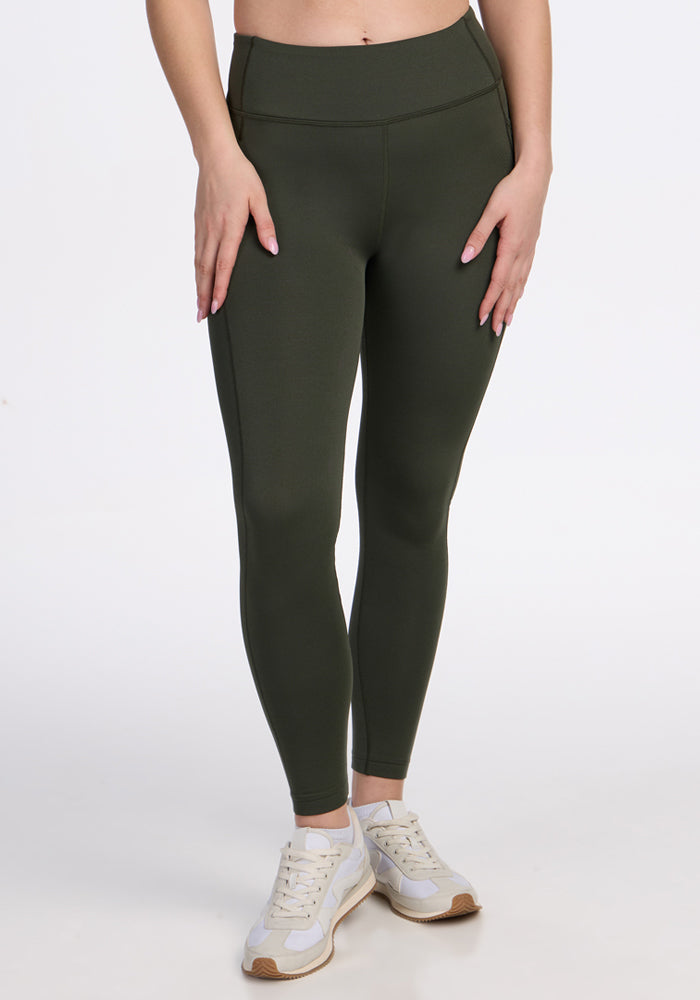 A person wearing Woolx Merino Flex® McKenna Petite - Everleaf high-waisted dark green leggings with pockets and white sneakers stands against a plain white background, showing only their lower body with hands resting on their thighs.