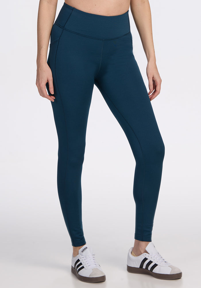 A person wearing Woolx Merino Flex® McKenna Petite leggings in Majolica Blue stands against a plain white background, paired with white sneakers with black stripes. The image shows only the lower half of their body.