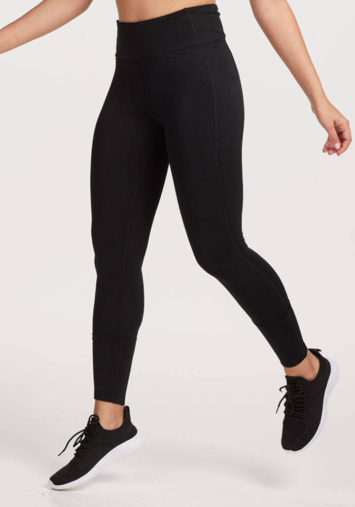 A person models Woolx Merino Flex® McKenna Petite leggings in black with athletic shoes, standing against a plain light backdrop. Only the lower body is visible, shown with one leg bent and arms slightly out.
