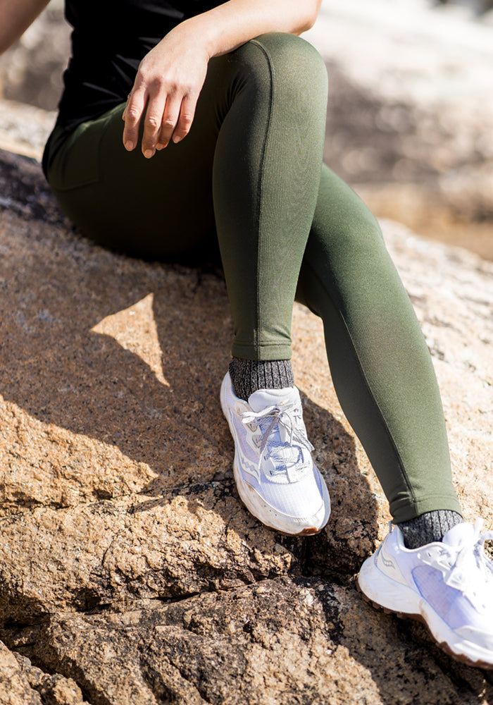 A person sits on a large rock outdoors, wearing Woolx Merino Flex® McKenna Pocket Leggings in Everleaf, paired with gray socks, white sneakers, and a black top, with their hand resting on their knee.