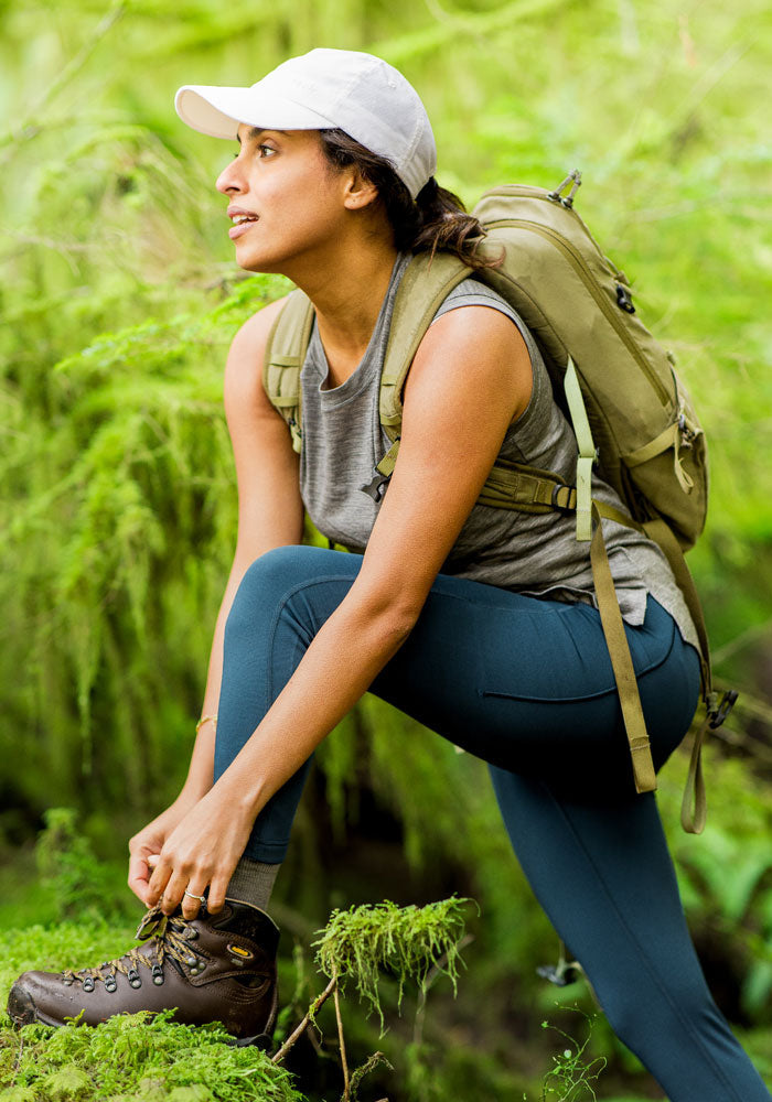 A woman in a white cap, gray tank top, and hiking boots kneels on a mossy rock in a lush forest, tying her shoe. She wears Woolx Merino Flex® McKenna Pocket Leggings in Majolica Blue and carries a beige backpack, gazing into the distance.