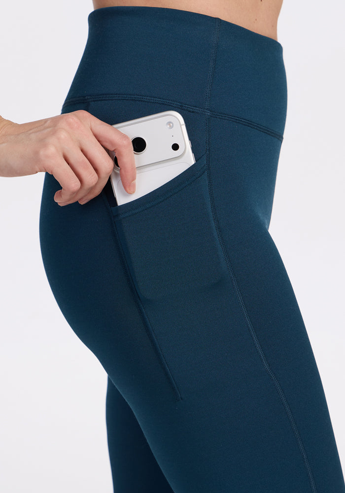 A person in Woolx Merino Flex® McKenna Pocket Leggings in Majolica Blue slides a white smartphone into a side pocket. The image is cropped to highlight the waist and upper leg area.