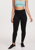 A woman stands against a white background wearing Woolx Merino Flex® McKenna Tall high-waisted black leggings with side pockets and a light blue sports bra. Her arms are relaxed at her sides, and her head is not visible.