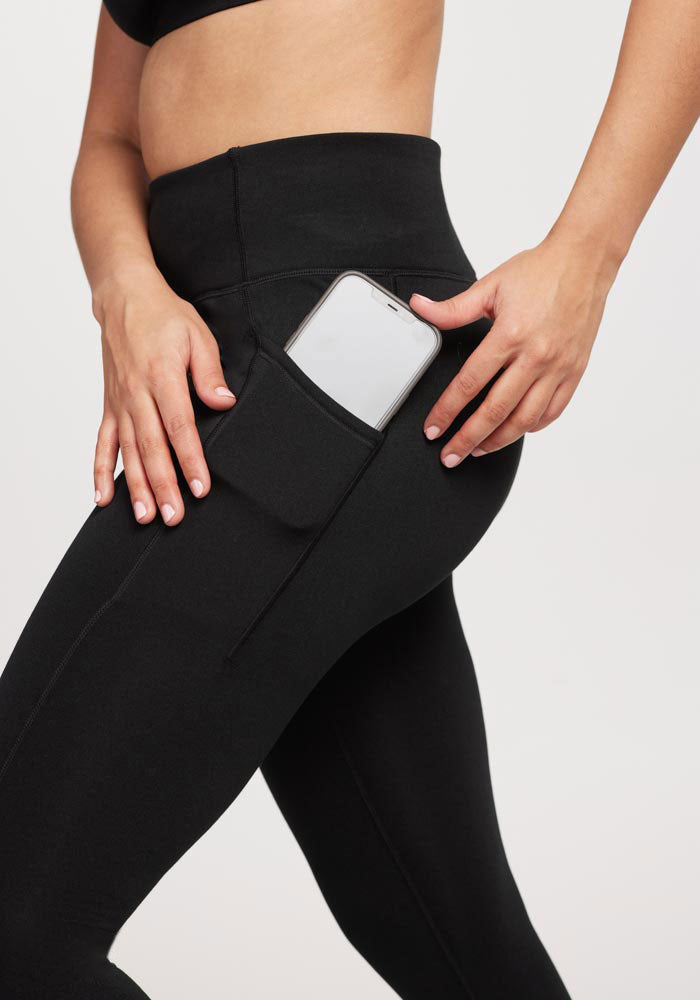A person wearing Woolx Merino Flex® McKenna Tall - Black leggings, shown from the waist down, places a smartphone into the sleek side pocket, highlighting convenient storage and the stylish design against a plain background.